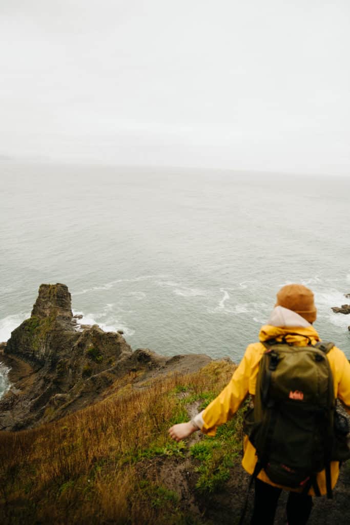 A girl in a yellow rain jacket is standing on a cliff overlooking the ocean at Oswald West State Park.