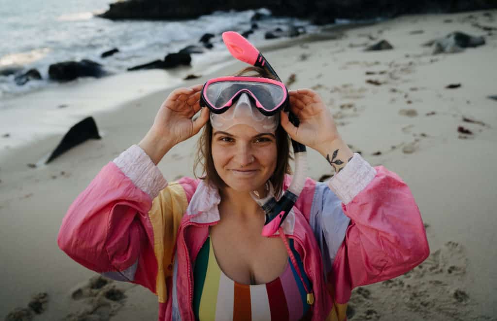 A girl is smiling on the beach at La Jolla Cove, while putting on a pink mask and snorkel.
