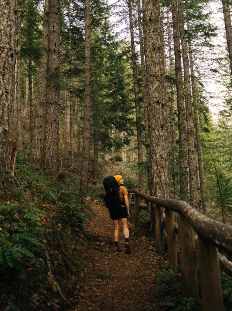 A girl is walking on the Lena Lake trail, wearing a black backpack. The trail is surrounded by trees.