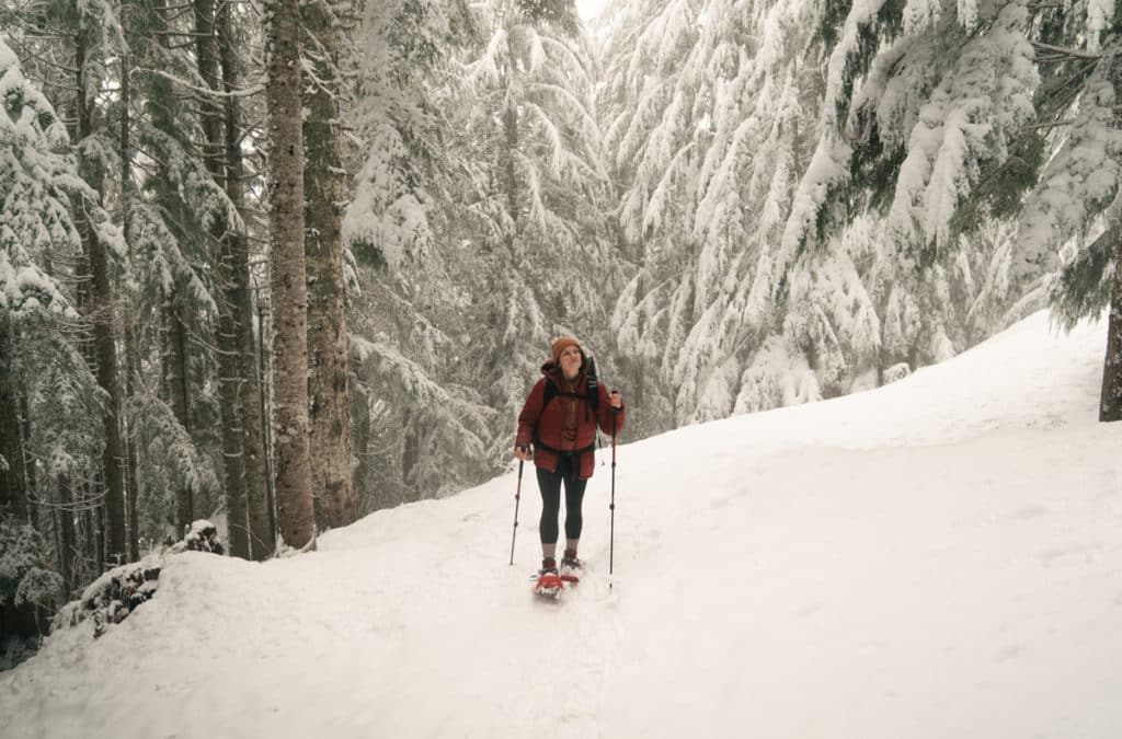 A girl wearing a red jacket is snowshoeing on the trail to Scenic Hot Springs, with snow on the ground and trees surrounding the trail.
