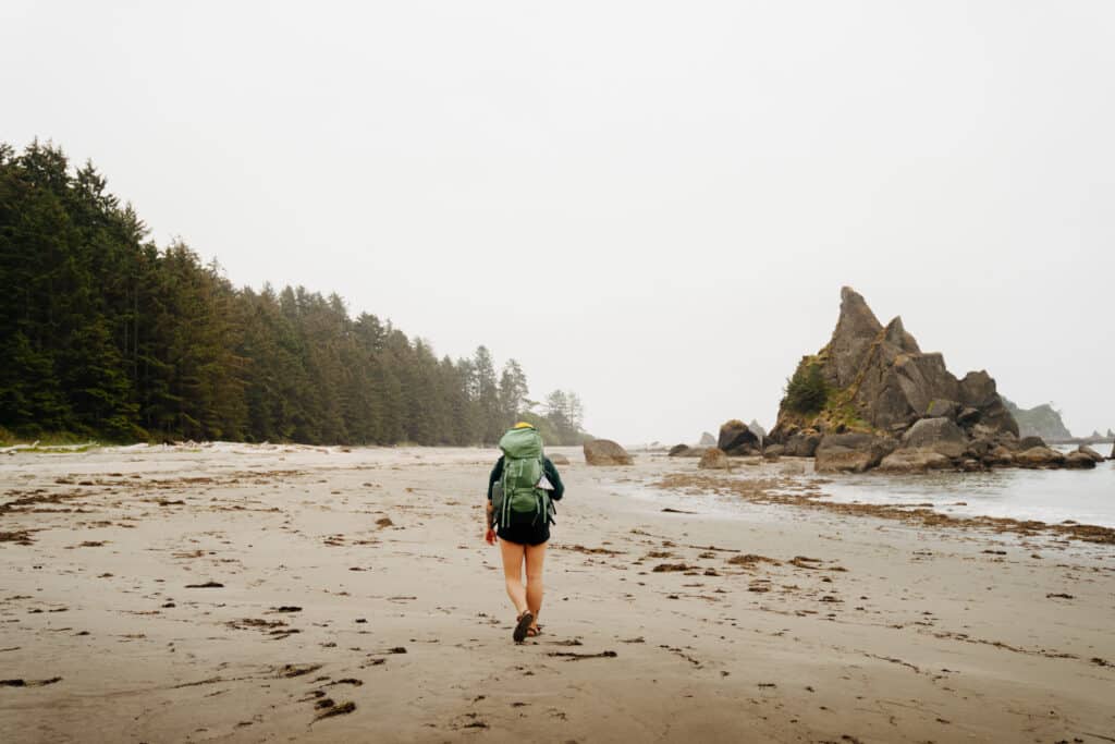 A girl with a green pack walking on the beach along the South Coast Wilderness trail in Olympic National Park.