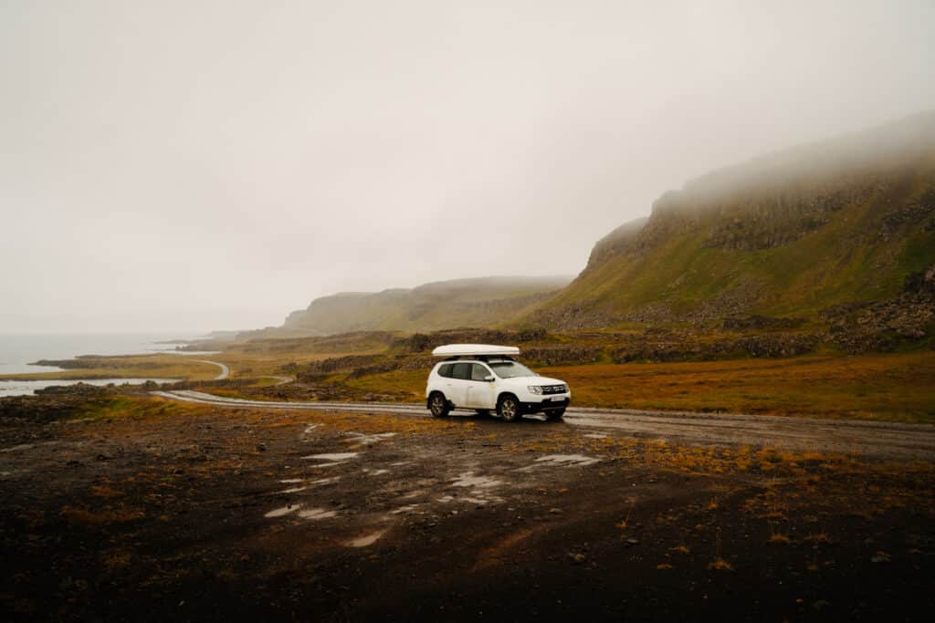 A white Dacia Duster driving through the Westfjords of Iceland, with mountains in the background.