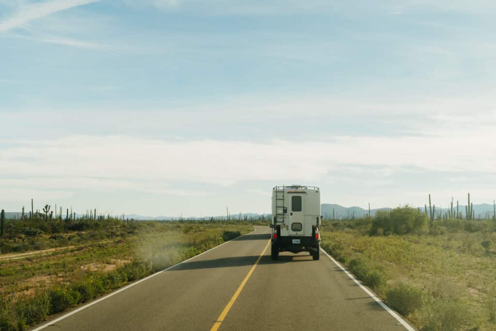 A truck camper driving to Baja, on a road surrounded by Saguaro cactuses.