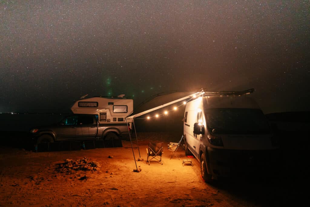 A van and a truck camper parked on a beach in Baja. It's night time, with stars in the sky and string lights illuminating the rigs.