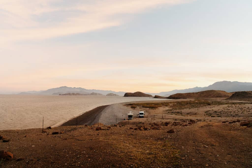 A van and a truck camper parked on a remote beach in Baja, with the ocean to the left and mountains in the backdrop.