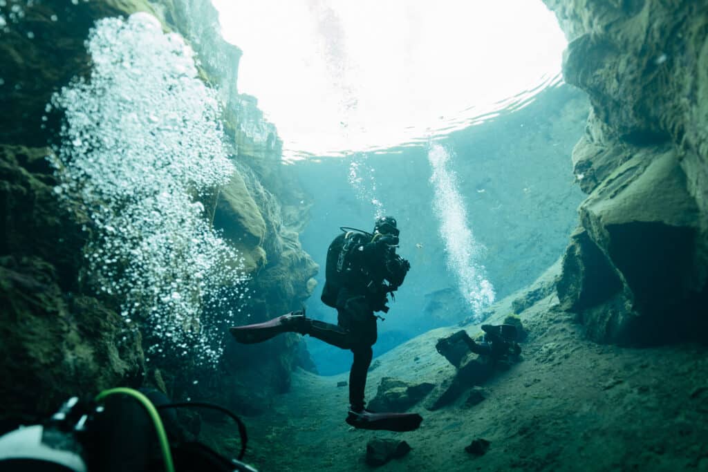 A scuba diver holding a camera and taking a photo underwater, surrounded by the rocky walls of the Silfra Fissure.